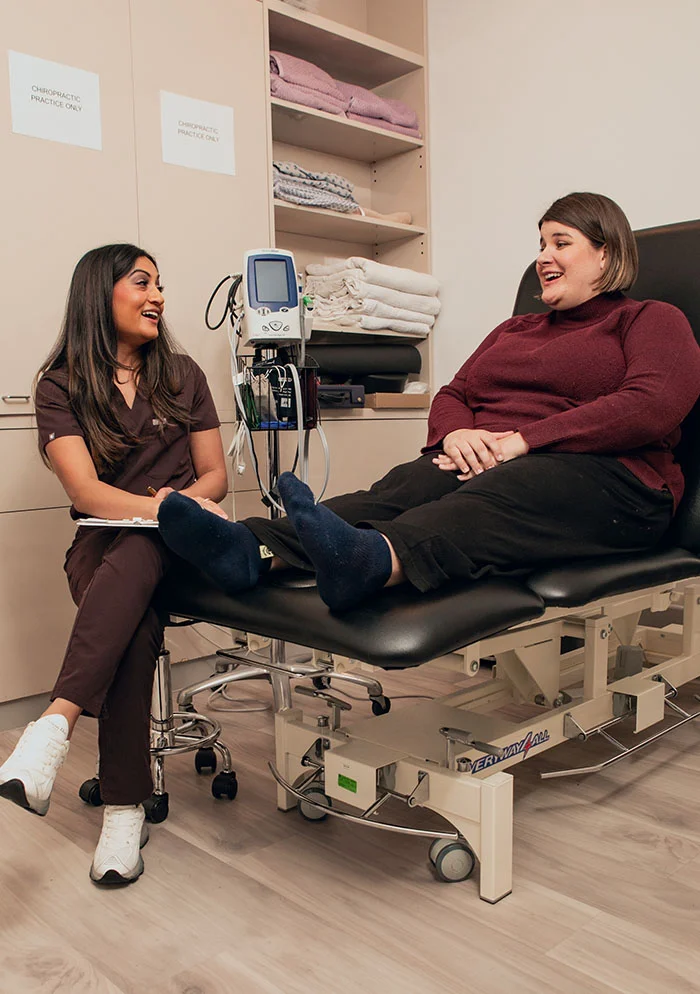 Dr. Ramani, a light-skinned woman in a brown scrub top and white sneakers, smiles at a second woman with light-brown hair seated on a black examination table. The second woman, wearing a maroon turtleneck sweater, is leaning back with her feet up. The background shows beige cabinets, medical supplies, and a medical device with a large screen. The setting is a medical clinic or physiotherapy office. - Tenax for Tendonitis Plantar Fascitis in New York, NY