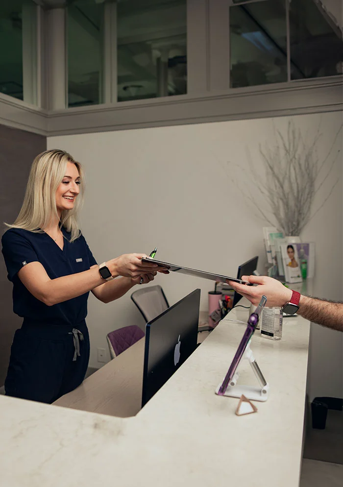 A friendly medical receptionist welcomes a patient in a modern and well-lit medical office. The receptionist, wearing a navy-blue scrub top, smiles warmly as she hands a clipboard to a patient at the reception desk. The clean and inviting atmosphere, with comfortable seating and modern decor, creates a positive and welcoming experience for patients. - Athletes Foot in New York, NY