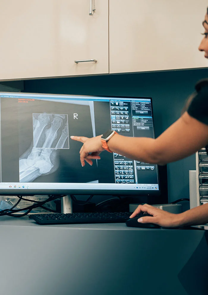 A medical professional with dark hair pulled back reviews a detailed foot X-ray on a computer monitor. Wearing a dark medical top, she points to a specific area on the screen, which also displays diagnostic software. The monitor sits on a gray desk with a keyboard and mouse. The background includes beige wall cabinets with metallic pulls and blue accents. - Bone Deformities in New York, NY