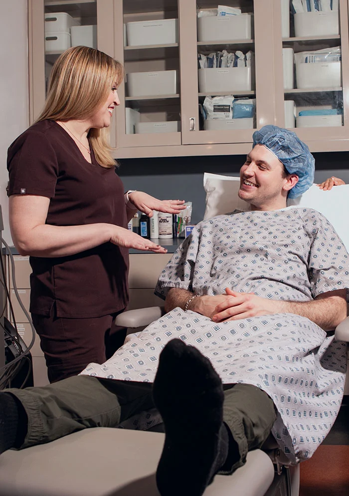 A healthcare provider in maroon scrubs stands beside a patient lying on a gray exam table. The patient, wearing a surgical gown and cap, smiles up at Dr. Koprince. - Cryotherapy Neuroma in New York, NY