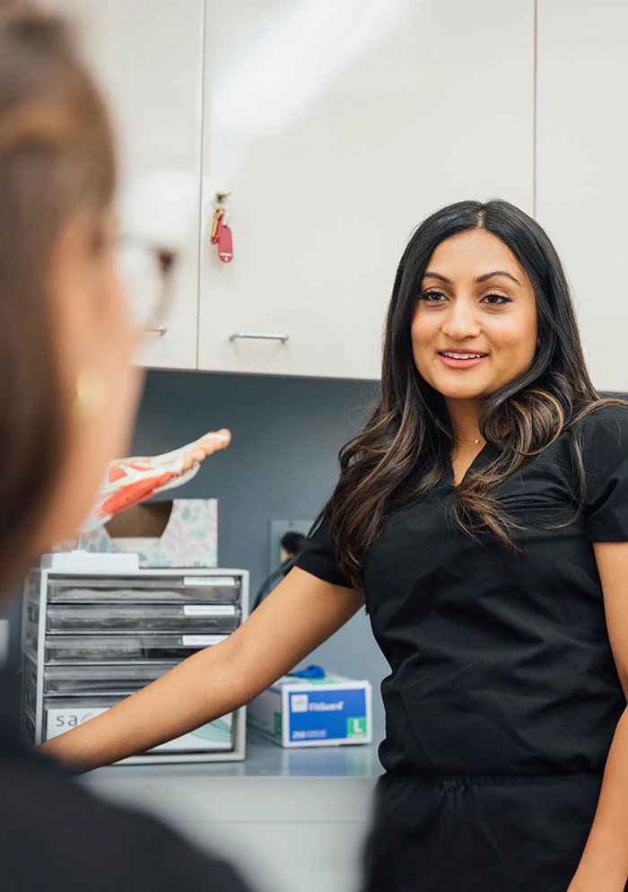 A close-up view of Dr. Mona Ramani, a woman with shoulder-length brown hair and a warm smile, in a professional setting. She is wearing a dark charcoal scrub top with a crew neckline, looking directly at the viewer with a pleasant expression. Behind her, a second woman with darker, out-of-focus hair is partially visible. The background includes white wall-mounted cabinets, a gray metal file cabinet, and various medical supplies, suggesting a healthcare office. - Flat Feet in New York, NY