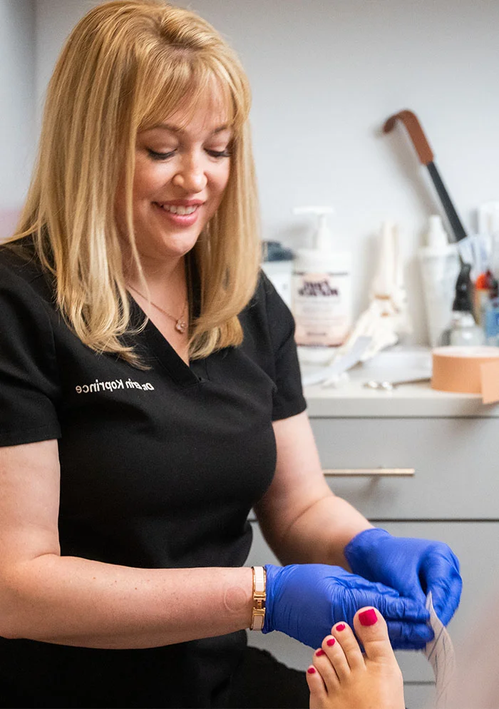 Dr. Erin Koprince, a podiatrist with shoulder-length blonde hair, smiles as she examines a patient’s foot. She wears a navy-blue or black short-sleeved top, light-blue nitrile gloves, and jewelry on her right wrist. The patient’s foot, with bright pink nail polish, is held for examination. The background features a light gray/white wall, medical equipment, and anatomical models, illuminated by bright, even lighting. - Foreign Body Removal in New York, NY