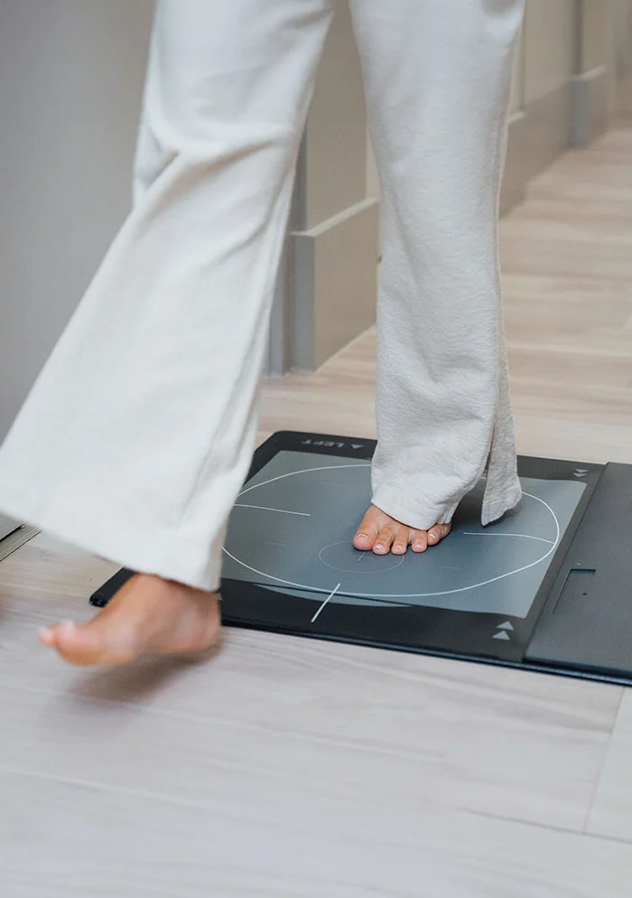 Full indoor shot of a person’s bare feet on a gray rectangular platform. The platform, part of a medical device, has a circular gray area for the feet. The person is wearing white pants, and their feet rest on the gray area. The platform is dark gray/black, placed on a light wood-style floor. The background features a light beige or cream-colored wall. - Gait Analysis in New York, NY