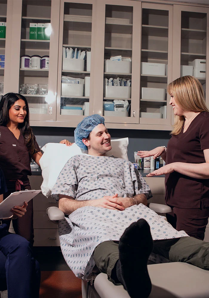 A healthcare provider in maroon scrubs stands beside a patient lying on a gray exam table. The patient, wearing a surgical gown and cap, smiles up at Dr. Koprince and Dr. Ramani is seen smiling in he background. - Hammer Toes in New York, NY