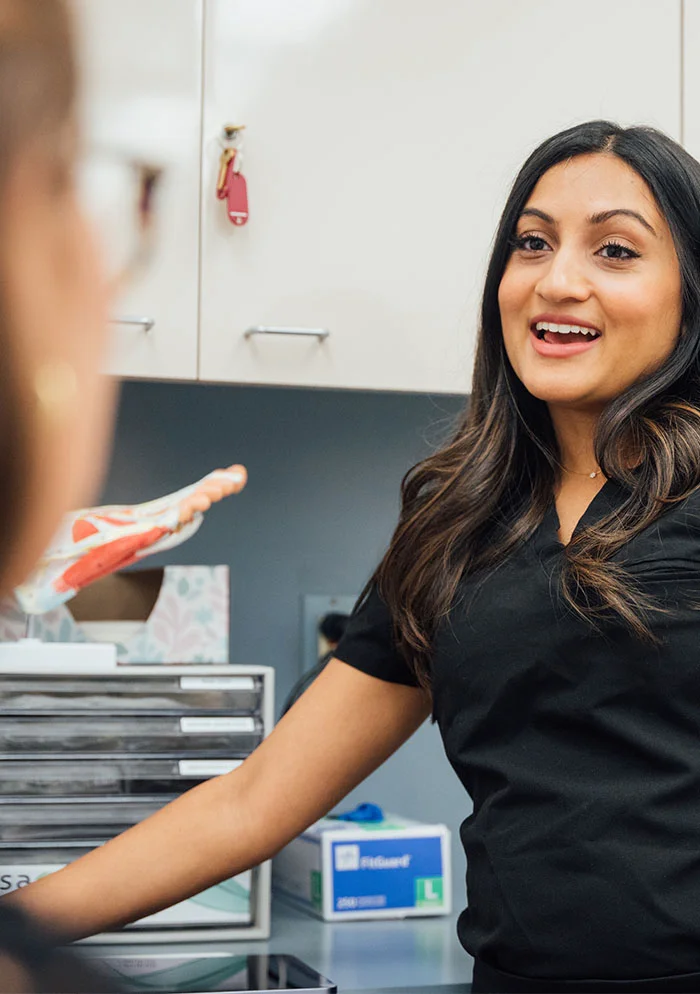 Dr. Ramani, with shoulder-length dark brown hair, smiles warmly in dark-colored medical scrubs. She engages directly with the viewer while her left arm reaches out. A portion of another person’s face and upper torso is seen in the foreground. The background shows white cabinets, lab equipment, and medical supplies on a table, with bright, even lighting. - Ingrown Toenail Treatment in New York, NY