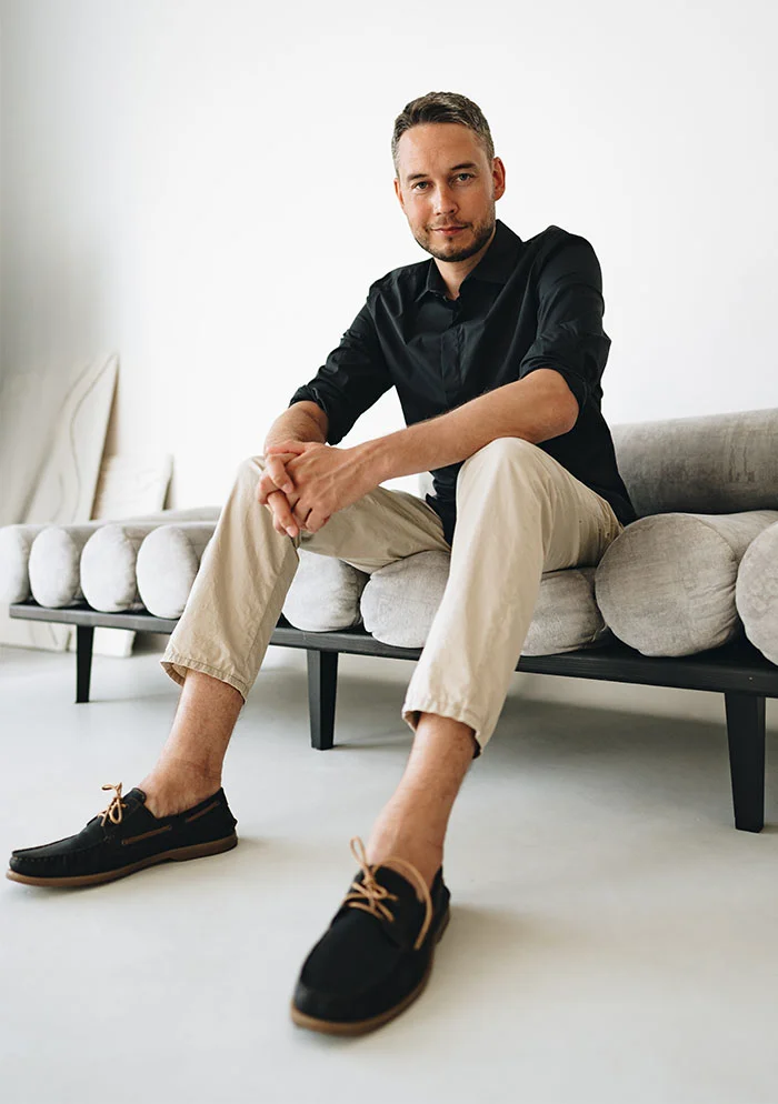 A light-skinned man sits on a modern, light gray, modular sofa, dressed in a black long-sleeved shirt and beige trousers. He looks directly at the camera, with relaxed posture. The plain white wall and light gray floor create a clean, minimalistic background. - Plantar Faceilitis in New York, NY