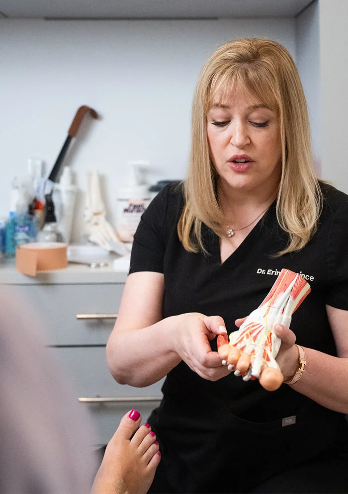 Dr. Koprince, a light-skinned woman with shoulder-length blonde hair, holds a 3D anatomical model of a foot with detailed muscles and bones. She is wearing a black, short-sleeved top and a necklace, with a focused expression. In the foreground, a person’s bare foot with pinkish-purple toenails is partially visible. The background features a white-walled room with beige cabinets and medical supplies, indicating a healthcare setting. - Shockwave Therapy in New York, NY