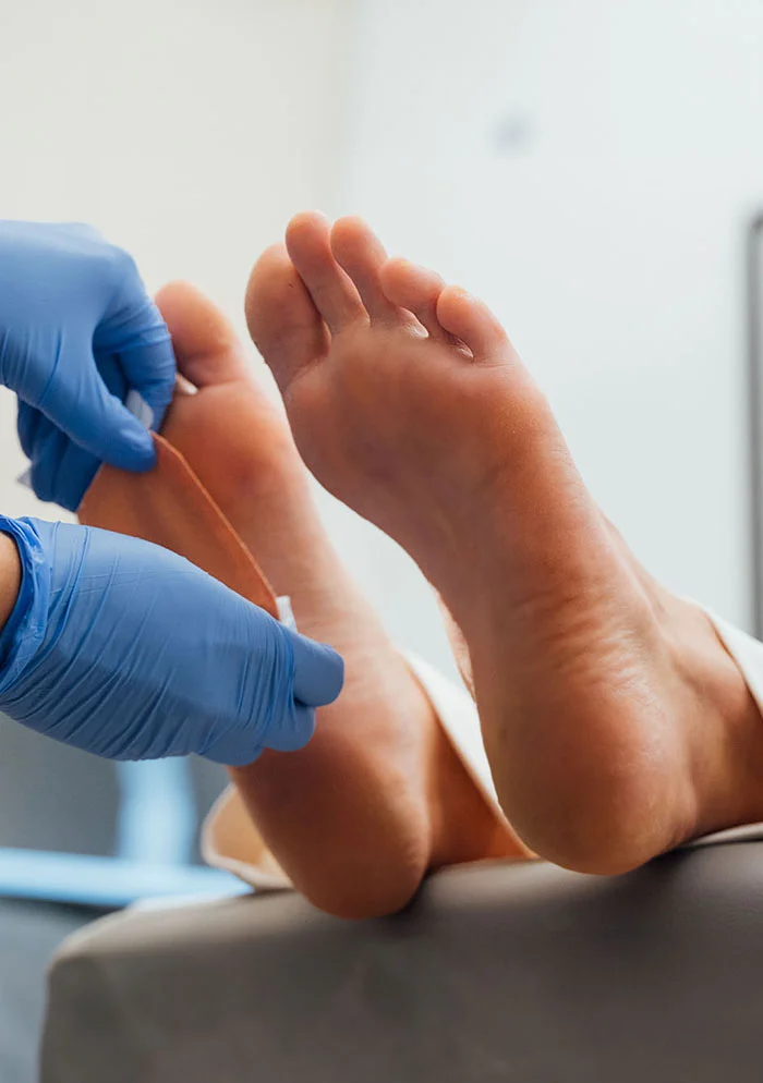 Close up shot of person's light brownish-tan foot rests on a light beige examination table while a medical professional in light-blue nitrile gloves examines it. The professional's hands and arms are visible as they apply a small probe or measuring device to the foot's sole. The background features a light gray/white wall with a framed image, suggesting a medical examination setting. - Skin in New York, NY