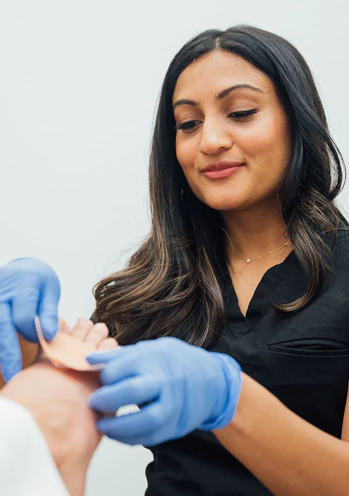 Dr. Ramani, wearing a dark gray scrub top and blue gloves, carefully applies a medical dressing to a person's foot. The background is a plain light beige wall. - Sprain and Strains in New York, NY