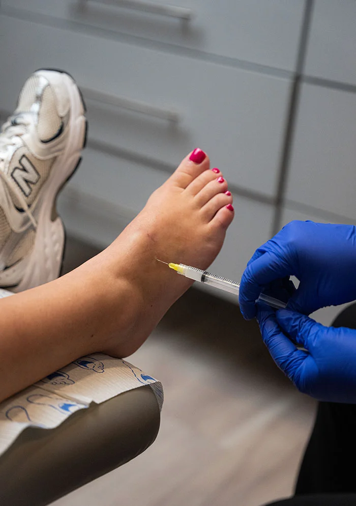 A bare foot with pink nail polish is angled up while a medical professional, wearing blue gloves, holds a syringe above the ankle. The person is seated in a medical chair with a paper pad under their leg, and white sneakers are visible. The background shows blurred gray cabinetry. - Stemcell Therapy in New York, NY